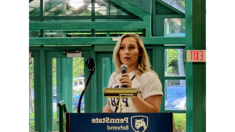 A person stands at a podium giving a speech, the podium has a banner that reads Penn State Behrend